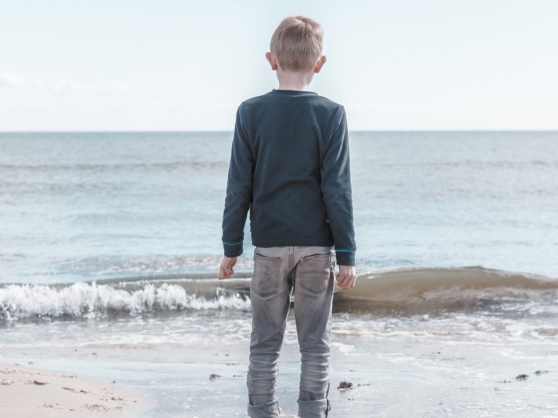 Blond boy in jeans and a blue sweater standing barefoot in the ocean, looking nervous