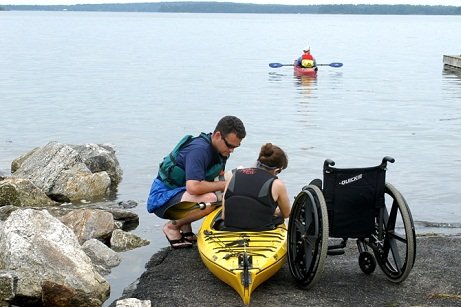 A man helps a woman into a kayak while her wheelchair sits nearby.