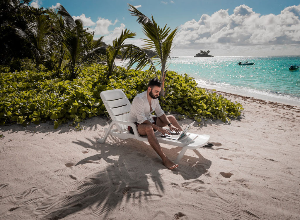 A man sitting on a deckchair at the beach and working on a laptop with the sea next to him